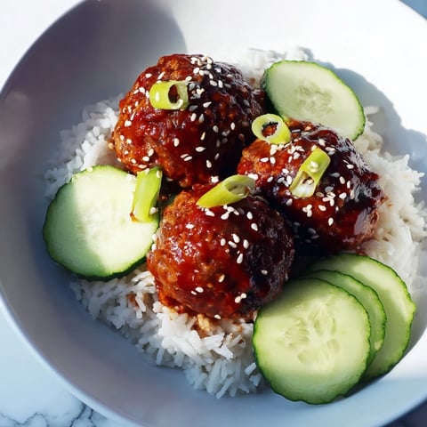 Steaming rice and glistening teriyaki meatball bowls, garnished with cucumber and sesame seeds, ready to eat.