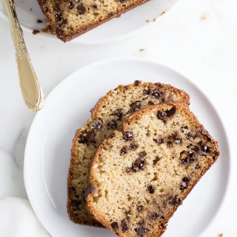 A freshly baked loaf of banana bread with chocolate chips sits on a wooden cutting board, ready to slice.  