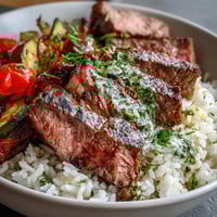 Freshly grilled steak slices rest atop fluffy white rice, colorful roasted vegetables, and a generous drizzle of bright green chimichurri sauce in this vibrant Grilled Steak Bowl.